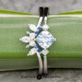 The "Selene" ring in 18k white gold with a 0.98-carat Montana sapphire features a large round center stone, accented by blue and clear marquise and round stones with a Double Band Jacket, displayed on a green background.