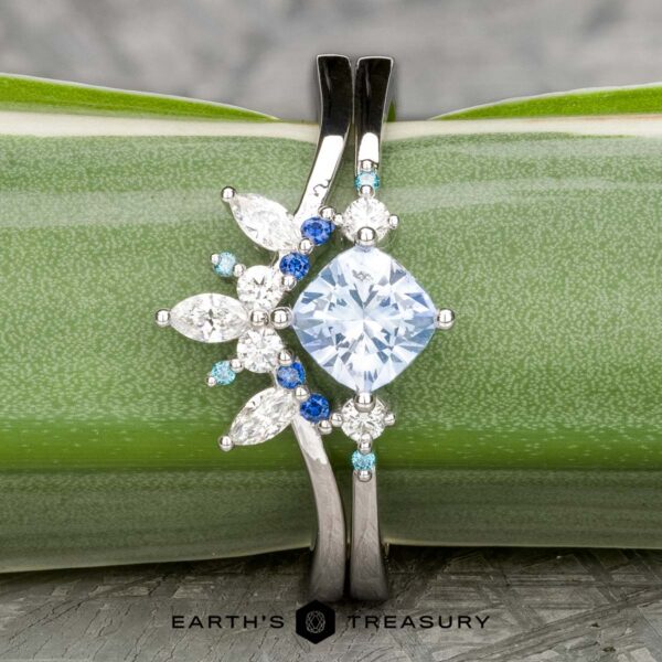 The "Selene" ring in 18k white gold with 0.98-carat Montana sapp The "Selene" ring in 18k white gold with a 0.98-carat Montana sapphire features a large round center stone, accented by blue and clear marquise and round stones with a Double Band Jacket, displayed on a green background.