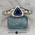 The “Foy, Andrew” white gold Corvus Ring, featuring a trilliant-cut blue sapphire center and round diamond accents, is displayed on a rough blue gemstone with the “Earth’s Treasury” logo below.