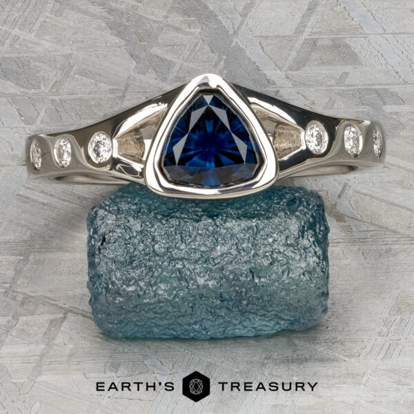 Foy, Andrew The “Foy, Andrew” white gold Corvus Ring, featuring a trilliant-cut blue sapphire center and round diamond accents, is displayed on a rough blue gemstone with the “Earth’s Treasury” logo below.