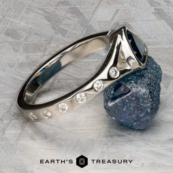 Foy, Andrew The Foy, Andrew ring, featuring a blue gemstone and five small diamonds, is displayed leaning on a rough blue stone atop a textured metallic surface with the Earth’s Treasury logo below.
