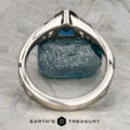 A close-up of the Foy, Andrew ring—a silver band with a blue gemstone—viewed from the side on a textured gray surface, with the "Earth's Treasury" logo beneath.