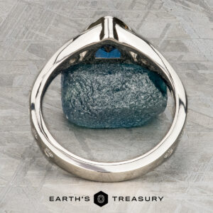 A close-up of the Foy, Andrew ring—a silver band with a blue gemstone—viewed from the side on a textured gray surface, with the "Earth's Treasury" logo beneath.