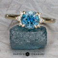 A Gaines, John (Jeremy) ring with a round blue gemstone and two small clear stones on each side rests on a rough blue crystal, displayed on a textured gray surface.