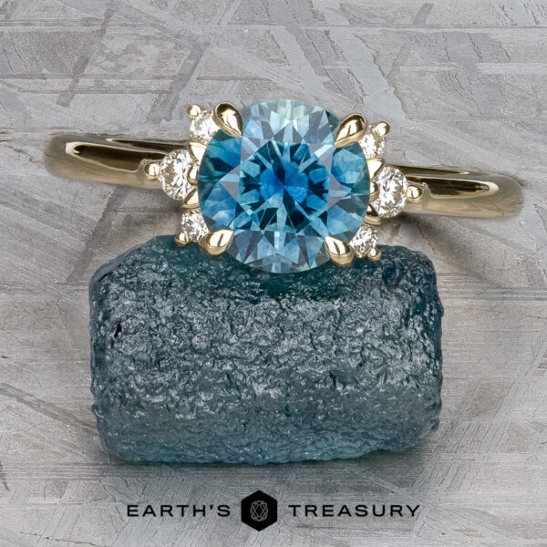 The "Lipara" ring in 14k yellow gold with 1.60-carat Montana sapphire A ring with a round blue gemstone and two small clear stones on each side rests on a rough blue crystal, displayed on a textured gray surface.