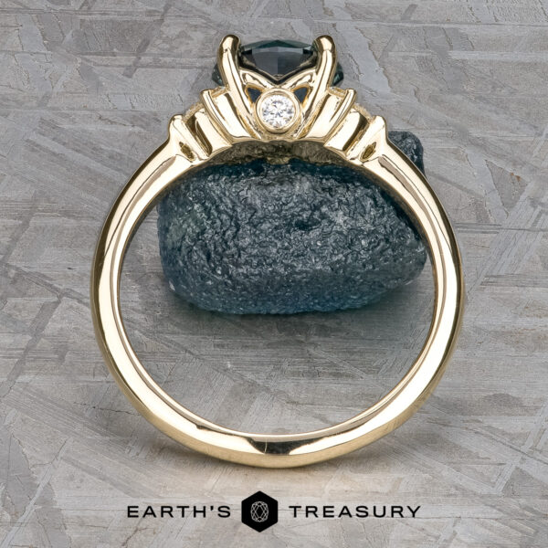The "Lipara" ring in 14k yellow gold with 1.60-carat Montana sapphire The gold Lipara Ring with a round white gemstone in a decorative setting is displayed upright on textured gray, dark stone behind, Earth’s Treasury logo below.