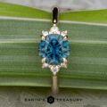 A gold Gaines, John (Jeremy) pendant with a round blue gemstone and six small diamonds, shown on a green leaf next to the Lipara Ring and the Earth's Treasury logo below.