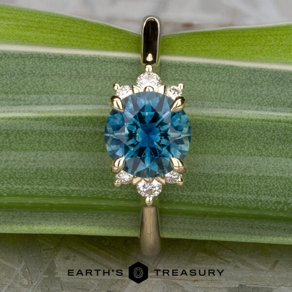 A gold Gaines, John (Jeremy) pendant with a round blue gemstone and six small diamonds, shown on a green leaf next to the Lipara Ring and the Earth's Treasury logo below.