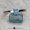 A "Narcissus" ring in 14K white gold with a 0.82-carat Montana sapphire rests on a rough blue stone, with "EARTH'S TREASURY" displayed below.