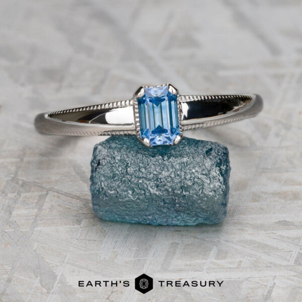 The "Narcissus" ring in 14K white gold with 0.82-carat Montana sapphire A "Narcissus" ring in 14K white gold with a 0.82-carat Montana sapphire rests on a rough blue stone, with "EARTH'S TREASURY" displayed below.