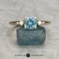 The "Selene" ring in 14K yellow gold, featuring a 1.34-carat cushion-cut Montana sapphire and two diamonds, is shown resting on a blue stone with the Earth's Treasury logo below.