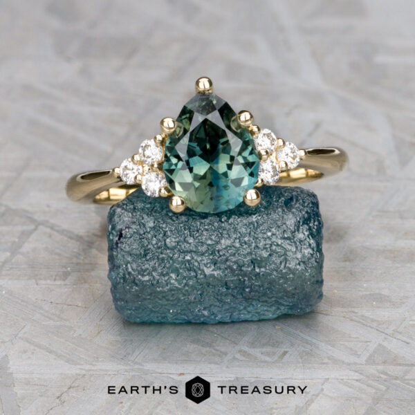 The "Kelli" ring in 14k yellow gold with 1.43-carat Australian s The "Kelli" ring in 14k yellow gold, set with a 1.43-carat Australian sapphire and accented by diamonds, is elegantly displayed on a rough blue-green stone.