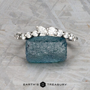 The "Ilana" band in platinum, featuring round and pear-shaped diamonds, is displayed on a rough blue gemstone with a textured gray background.