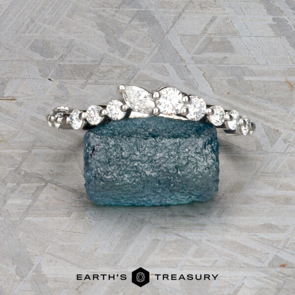 The "Ilana" band in platinum The "Ilana" band in platinum, featuring round and pear-shaped diamonds, is displayed on a rough blue gemstone with a textured gray background.