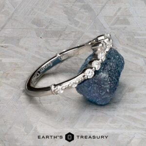 The "Ilana" band in platinum rests on a rough blue gemstone atop a textured gray surface, with the "Earth's Treasury" logo below.