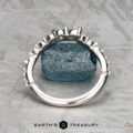A platinum "Ilana" band ring with prong settings is displayed upright, encircling a rough blue gemstone on a textured gray surface. The "Earth's Treasury" logo and text appear at the bottom.
