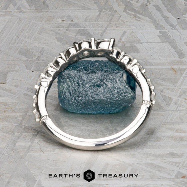 The "Ilana" band in platinum A platinum "Ilana" band ring with prong settings is displayed upright, encircling a rough blue gemstone on a textured gray surface. The "Earth's Treasury" logo and text appear at the bottom.