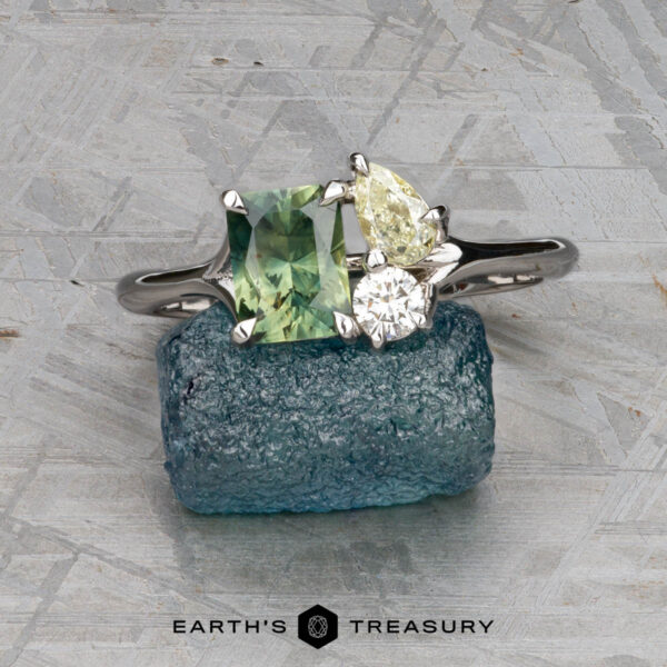 The "Moirae" ring in 14K white gold with 1.05-carat Australian ssapphire The "Moirae" ring in 14k white gold, featuring a 1.05-carat Australian sapphire, rests atop a rough blue stone on a textured gray surface. The Earth's Treasury logo is shown at the bottom.