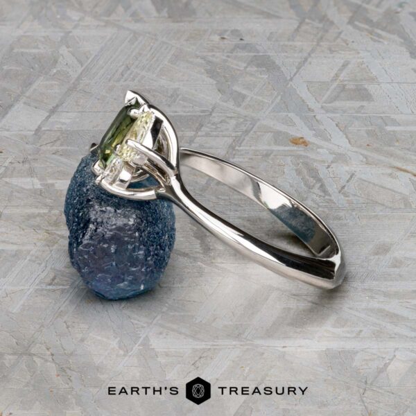 The "Moirae" ring in 14K white gold with 1.05-carat Australian ssapphire The "Moirae" ring in 14k white gold, featuring a 1.05-carat Australian sapphire, rests atop a rough blue stone on a textured gray surface. The Earth's Treasury logo is shown at the bottom.