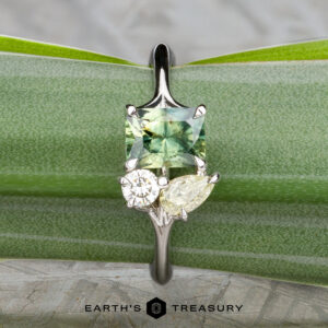 The "Moirae" ring in 14k white gold, featuring a 1.05-carat Australian sapphire, rests atop a rough blue stone on a textured gray surface. The Earth's Treasury logo is shown at the bottom.