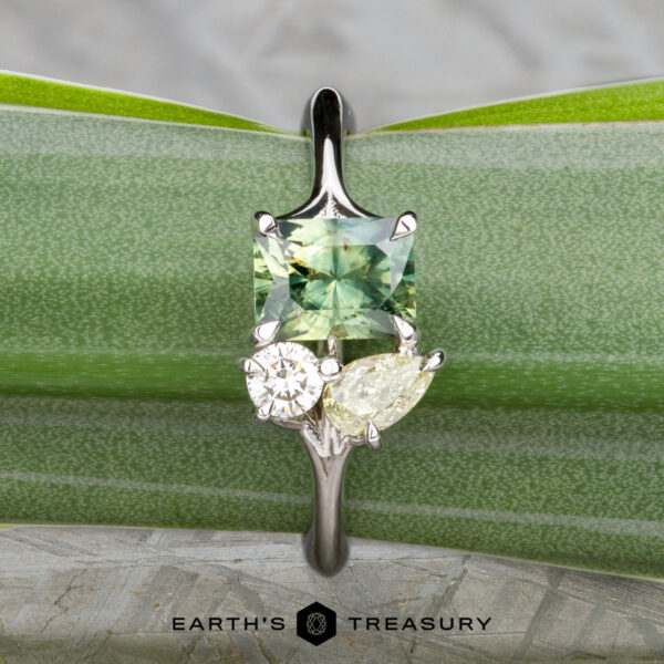 The "Moirae" ring in 14K white gold with 1.05-carat Australian ssapphire The "Moirae" ring in 14k white gold, featuring a 1.05-carat Australian sapphire, rests atop a rough blue stone on a textured gray surface. The Earth's Treasury logo is shown at the bottom.