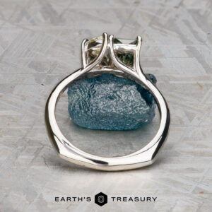 The "Moirae" ring in 14k white gold, featuring a 1.05-carat Australian sapphire, rests atop a rough blue stone on a textured gray surface. The Earth's Treasury logo is shown at the bottom.
