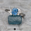 The Deluxe "Nebula" ring in 14k white gold features a 1.73-carat Montana sapphire and accent stones, displayed on a rough blue crystal with the Earth's Treasury logo at the bottom.