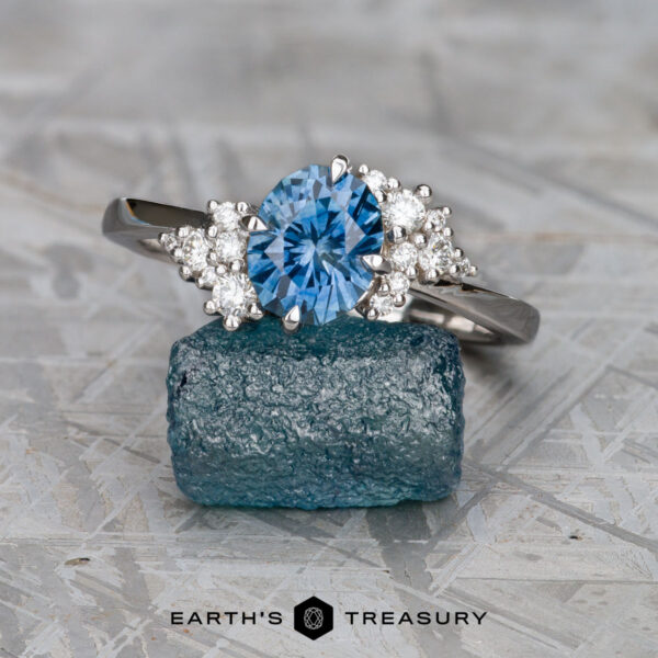 The Deluxe "Nebula" ring in 14k white gold with 1.73-carat Montana sapphire The Deluxe "Nebula" ring in 14k white gold features a 1.73-carat Montana sapphire and accent stones, displayed on a rough blue crystal with the Earth's Treasury logo at the bottom.