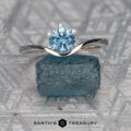 A "Ruenna" solitaire ring in 14k white gold with a 0.96-carat Montana sapphire rests on a rough blue crystal, displayed on a patterned gray surface with "Earth's Treasury" written below.