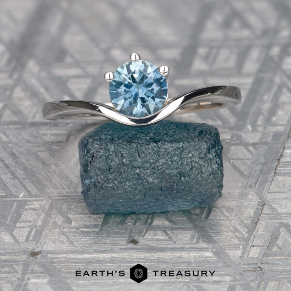 The "Ruenna" ring in 14k white gold with 0.96-carat Montana sapphire A "Ruenna" solitaire ring in 14k white gold with a 0.96-carat Montana sapphire rests on a rough blue crystal, displayed on a patterned gray surface with "Earth's Treasury" written below.