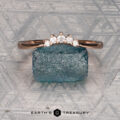 The "Avre" band in 14K rose gold, featuring five small diamonds, is balanced on a light blue textured stone against a patterned gray background. The Earth's Treasury logo is visible at the bottom.