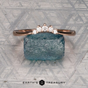 The "Avre" band in 14K rose gold, featuring five small diamonds, is balanced on a light blue textured stone against a patterned gray background. The Earth's Treasury logo is visible at the bottom.