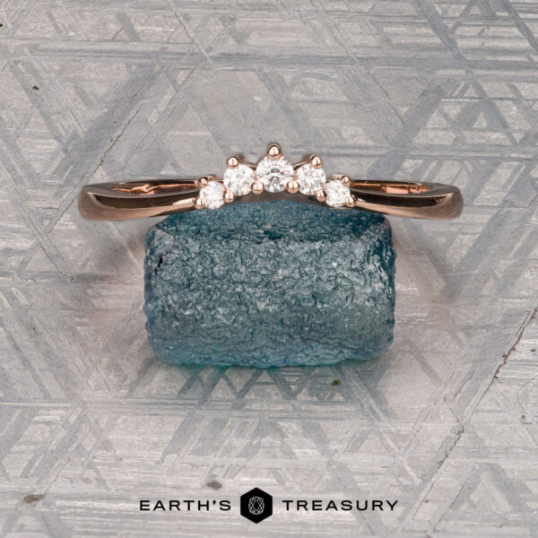 The "Avre" band in 14K rose gold The "Avre" band in 14K rose gold, featuring five small diamonds, is balanced on a light blue textured stone against a patterned gray background. The Earth's Treasury logo is visible at the bottom.