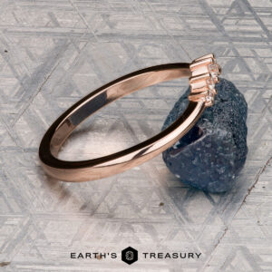 The "Avre" band in 14K rose gold, featuring five small diamonds, is balanced on a light blue textured stone against a patterned gray background. The Earth's Treasury logo is visible at the bottom.