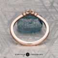 The "Avre" band in 14K rose gold, featuring five small diamonds, is balanced on a light blue textured stone against a patterned gray background. The Earth's Treasury logo is visible at the bottom.