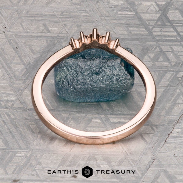 Gutierrez, Tomas 2 The "Avre" band in 14K rose gold, featuring five small diamonds, is balanced on a light blue textured stone against a patterned gray background. The Earth's Treasury logo is visible at the bottom.