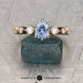 The "Solstice" ring in 14K yellow gold features a 0.90-carat Montana sapphire center stone with diamond accents, displayed on a blue-green mineral. The Earth’s Treasury logo appears below, evoking Solstice brilliance.