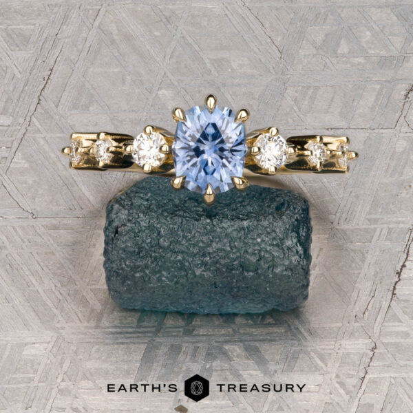 The "Solstice" ring in 14K yellow gold with 0.90-carat Montana sapphire The "Solstice" ring in 14K yellow gold features a 0.90-carat Montana sapphire center stone with diamond accents, displayed on a blue-green mineral. The Earth’s Treasury logo appears below, evoking Solstice brilliance.