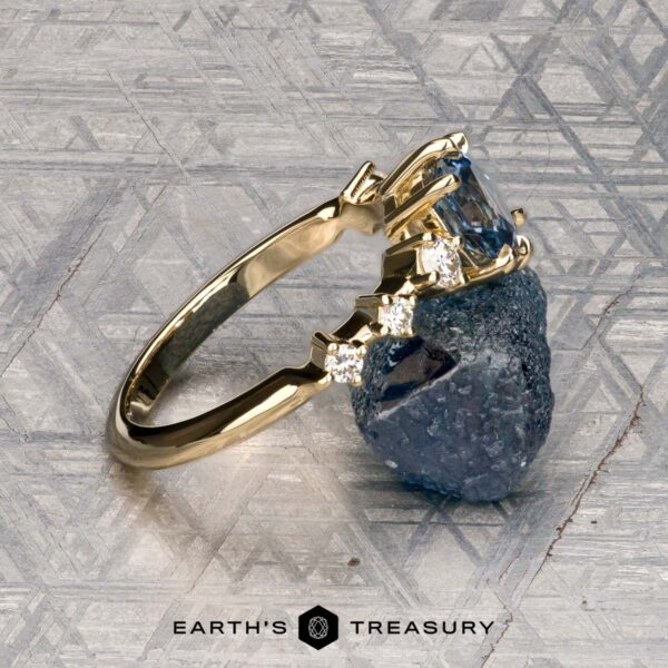 The "Solstice" ring in 14K yellow gold with 0.90-carat Montana sapphire The "Solstice" ring in 14K yellow gold features a 0.90-carat Montana sapphire center stone with diamond accents, displayed on a blue-green mineral. The Earth’s Treasury logo appears below, evoking Solstice brilliance.
