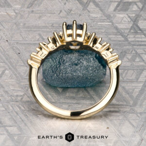 The "Solstice" ring in 14K yellow gold features a 0.90-carat Montana sapphire center stone with diamond accents, displayed on a blue-green mineral. The Earth’s Treasury logo appears below, evoking Solstice brilliance.