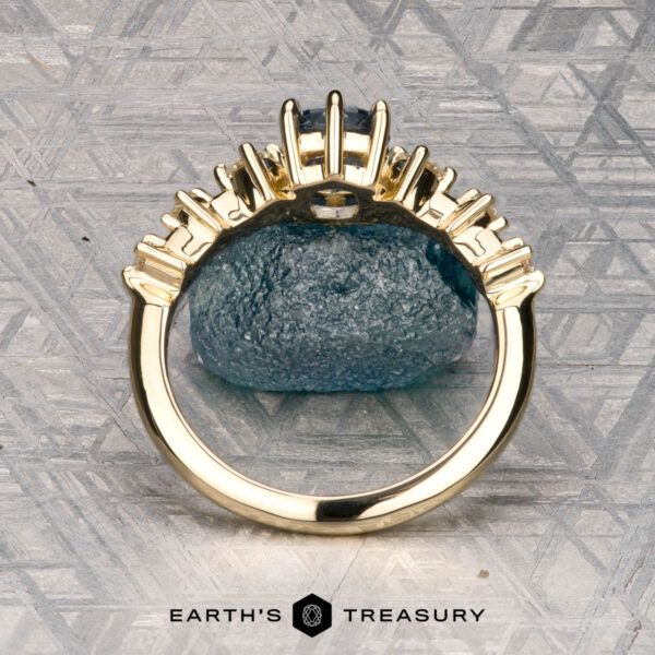 The "Solstice" ring in 14K yellow gold with 0.90-carat Montana sapphire The "Solstice" ring in 14K yellow gold features a 0.90-carat Montana sapphire center stone with diamond accents, displayed on a blue-green mineral. The Earth’s Treasury logo appears below, evoking Solstice brilliance.