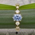 The "Solstice" ring in 14K yellow gold features a 0.90-carat Montana sapphire center stone with diamond accents, displayed on a blue-green mineral. The Earth’s Treasury logo appears below, evoking Solstice brilliance.