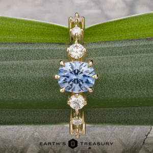 The "Solstice" ring in 14K yellow gold features a 0.90-carat Montana sapphire center stone with diamond accents, displayed on a blue-green mineral. The Earth’s Treasury logo appears below, evoking Solstice brilliance.