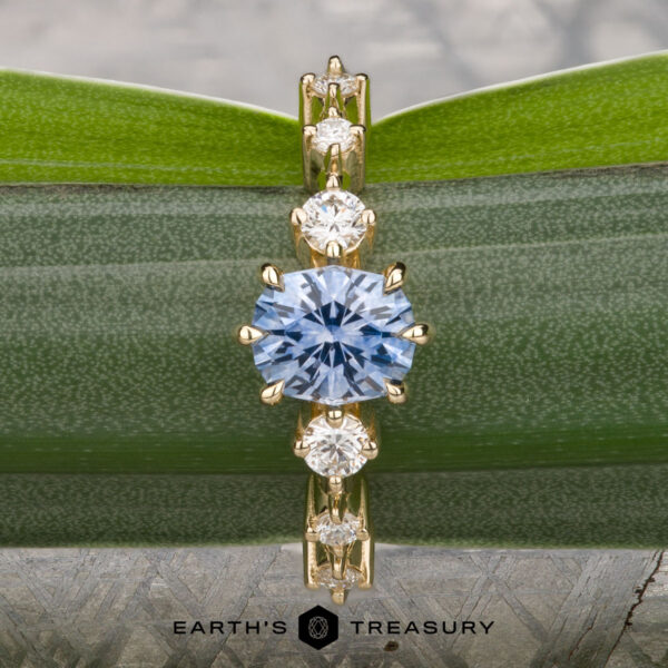 The "Solstice" ring in 14K yellow gold with 0.90-carat Montana sapphire The "Solstice" ring in 14K yellow gold features a 0.90-carat Montana sapphire center stone with diamond accents, displayed on a blue-green mineral. The Earth’s Treasury logo appears below, evoking Solstice brilliance.