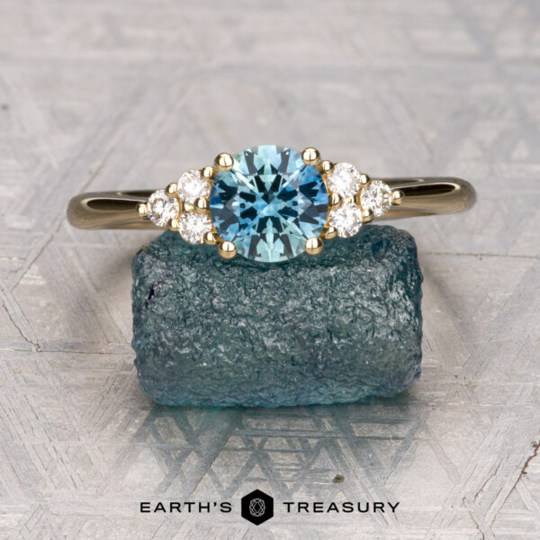 The "Kelli" ring in 14K yellow gold with 0.89-carat Montana sapphire The "Kelli" ring in 14K yellow gold, featuring a 0.89-carat Montana sapphire and white accent stones, is displayed on a rough blue mineral with the "Earth's Treasury" logo below.