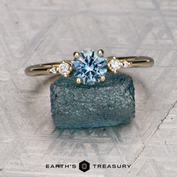 The "Selene" ring in 14K yellow gold with 0.98-carat Montana sapphire The "Selene" ring in 14K yellow gold, featuring a 0.98-carat Montana sapphire and two small diamonds, stands on a rough blue stone. The Earth's Treasury logo is visible at the bottom.