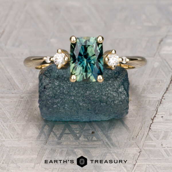The "Selene" ring in 14K yellow gold with 1.39-carat Australian sapphire The "Selene" ring in 14K yellow gold features a 1.39-carat Australian sapphire and two round diamonds, shown on a rough blue stone. The Earth's Treasury logo appears below.