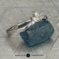 The "Alene" band in 14K white gold with an empty prong setting stands upright against a blue-green rough gemstone, with the Earth's Treasury logo visible at the bottom.
