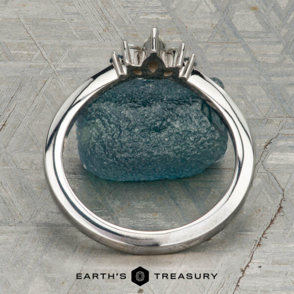 The "Alene" band in 14K white gold The "Alene" band in 14K white gold with an empty prong setting stands upright against a blue-green rough gemstone, with the Earth's Treasury logo visible at the bottom.