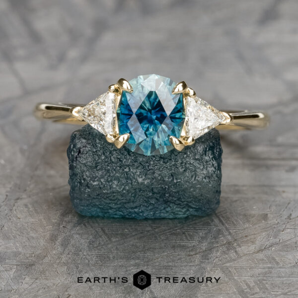 The Cathedral "Mavis" ring in 14K yellow gold with 1.58-carat Montana sapphire The Cathedral "Mavis" ring in 14K yellow gold features a 1.58-carat Montana sapphire flanked by two triangular diamonds, displayed on a rough blue crystal with the "Earth's Treasury" logo below.